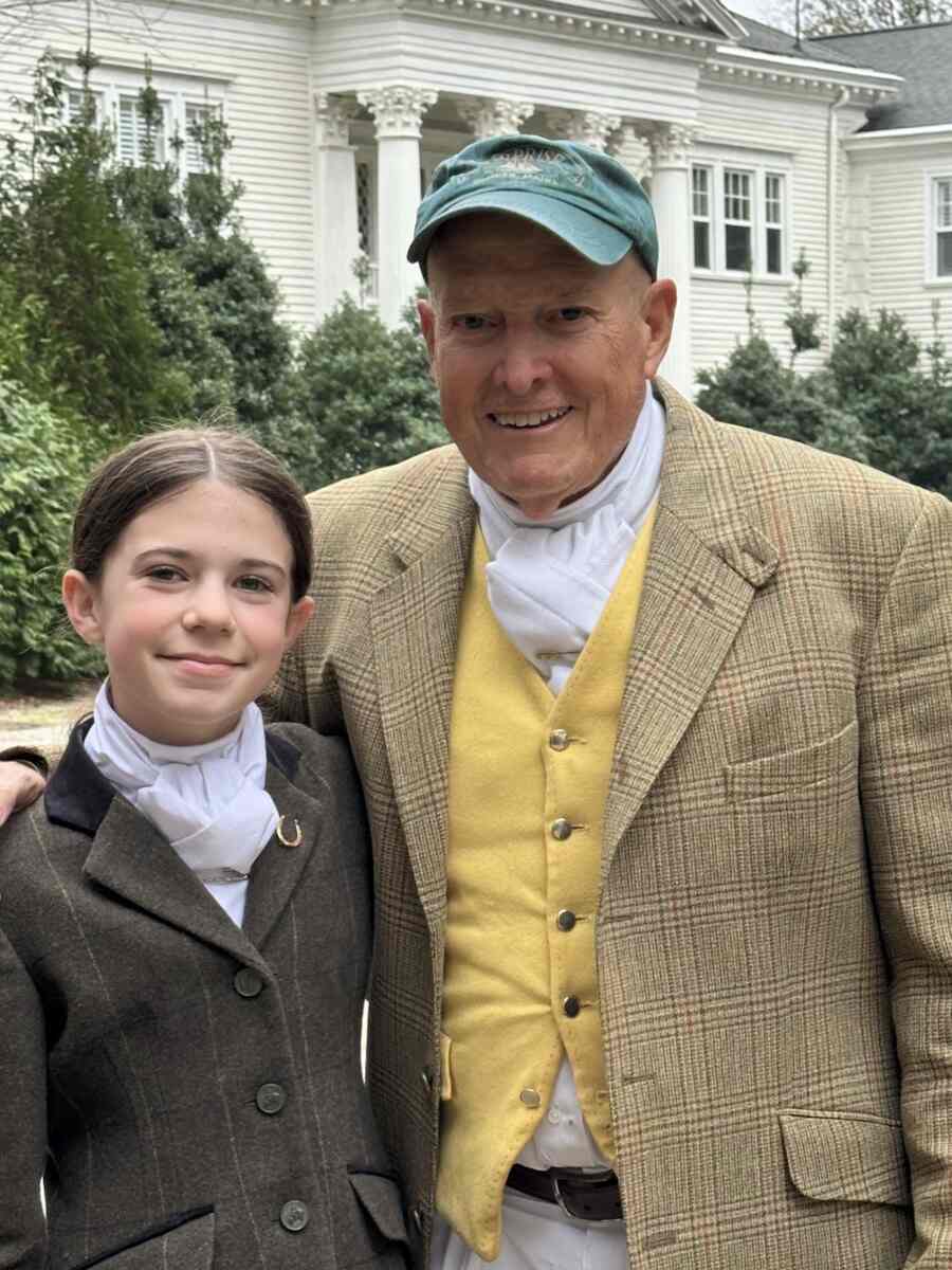 Man in tweed coat and yellow vest with girl in tweed coat, standing in front of white building.